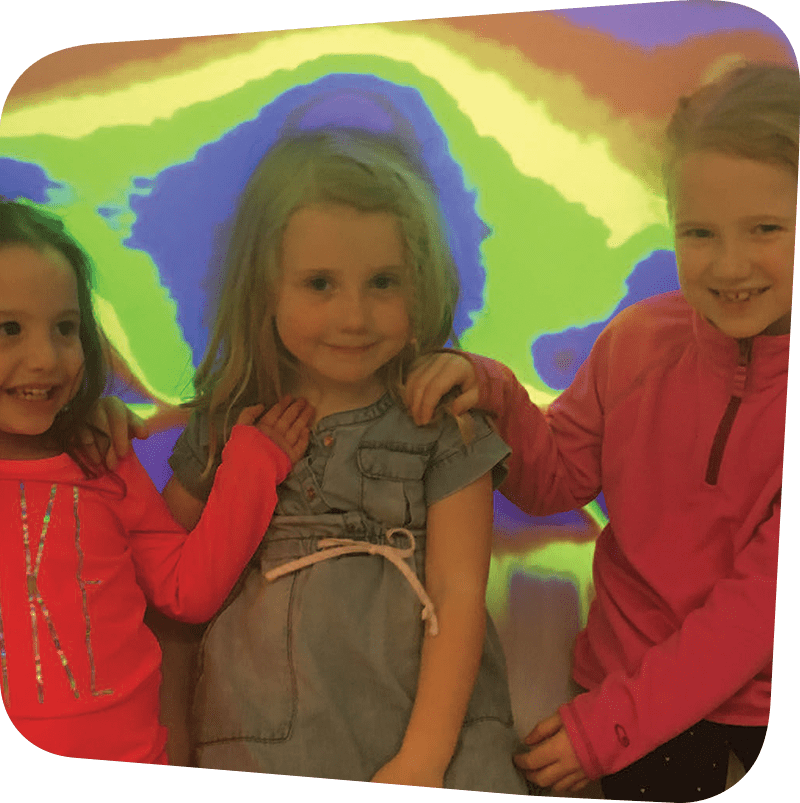 Three children smiling in front of a colorful wall.