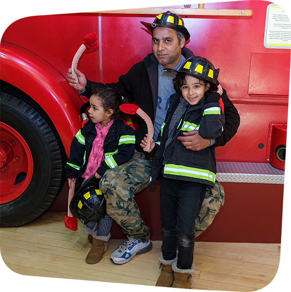 Children posing with parent in front of fire truck while wearing firefighter gear.