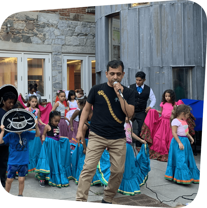 Man with microphone leading young dancers in Mexican traditional clothing through choreography