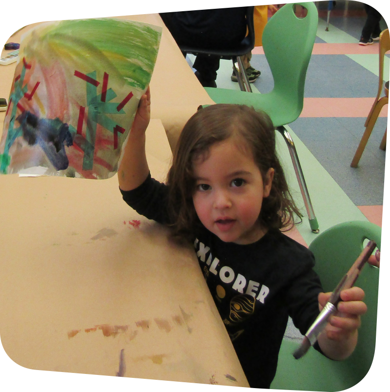 child displaying their art in one and and holding a paint brush in another while looking at the camera