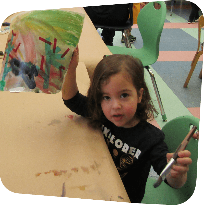child displaying their art in one and and holding a paint brush in another while looking at the camera