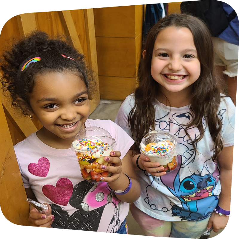 two girls smiling big grins and holding fruit cups they made