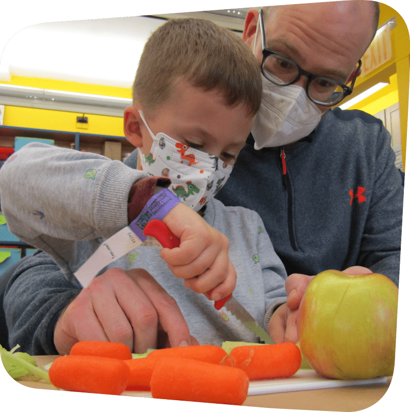 young boy cutting carrots with a serrated knife guided by his father