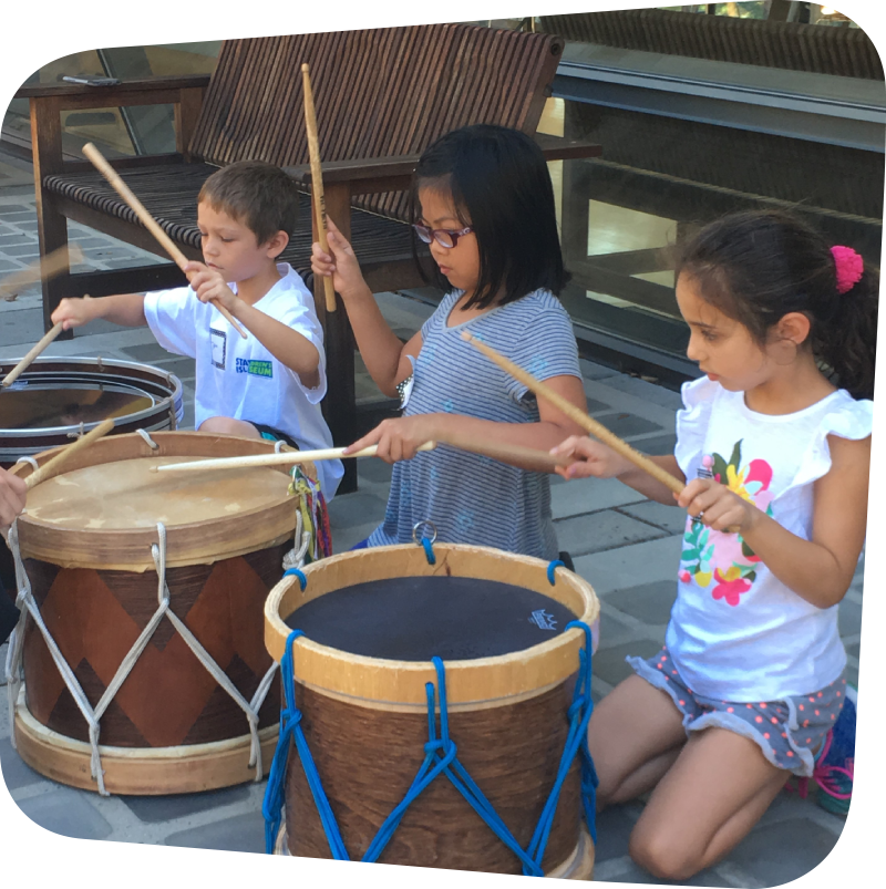 three children banging on large drums with drumsticks on the garden terrace