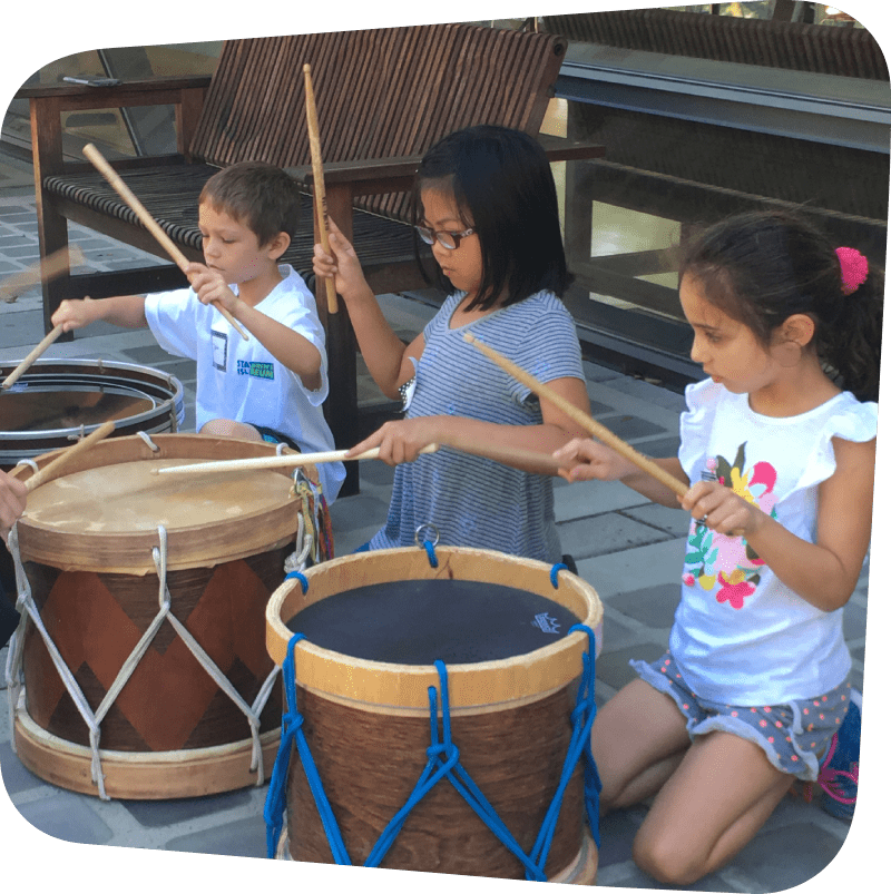 three children banging on large drums with drumsticks on the garden terrace