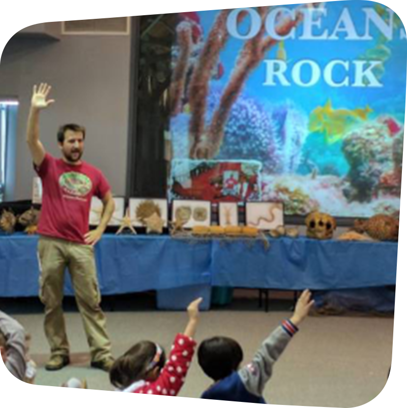 Man making a presentation to children with a large screen in the background showing an underwater scene of colorful rocks. In front of the screen is a long table displaying life-size taxidermy sea creatures.