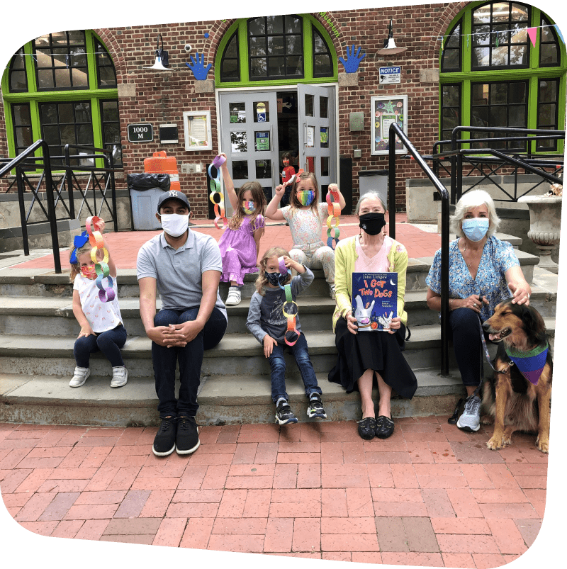 children, staff and volunteers sitting on the front steps of the museum with a dog displaying a children's book and crafts they've made