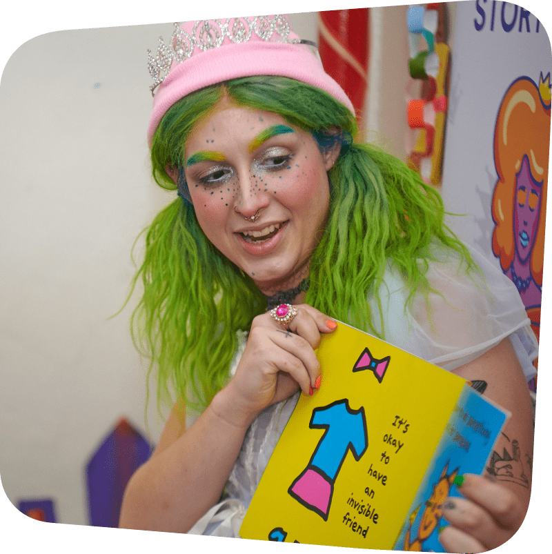 Someone dressed in their drag persona. They have green hair, with green matching eyebrows. They have on fun, playful makeup and a pink crown. They are holding a book.