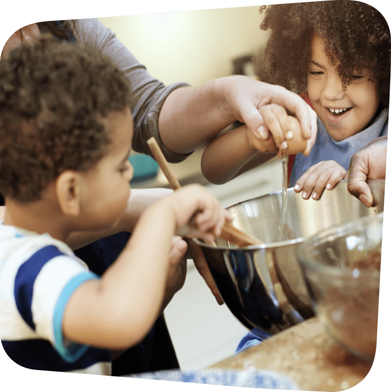 children with curly hair and light brown skin gleefully helping an adult add ingredients to a bowl for mixing