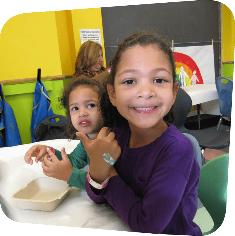 Two children in frame. One is wearing a purple long sleeve shirt and smiling at the camera. The other is wearing a teal long sleeve shirt and looking past the camera.