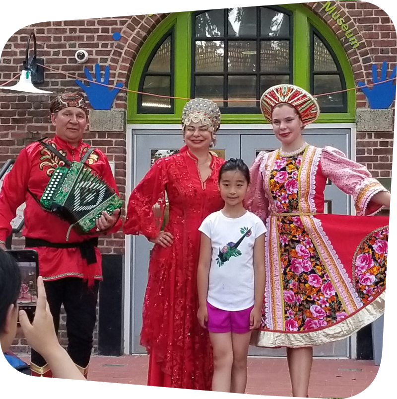 Performers wearing cultural attire posing for a picture with a smiling child in front of the children's museum.