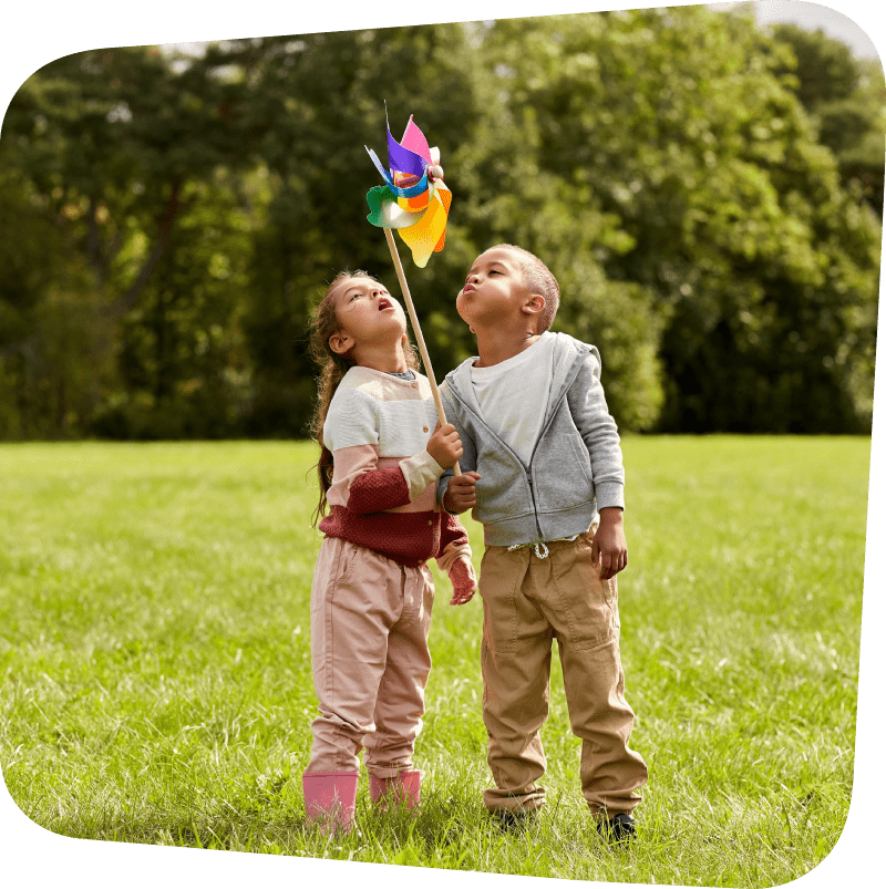Two children blowing on a pinwheel in the middle of a meadow with evergreen trees in the background