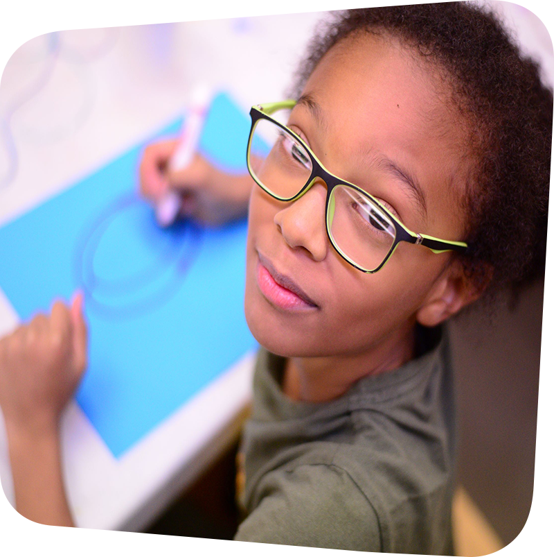 Child looking up at the camera from her work drawing on a blue sheet of paper with a red marker