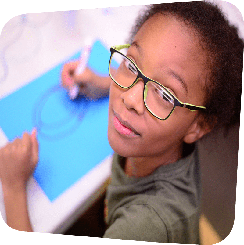 Child looking up at the camera from her work drawing on a blue sheet of paper with a red marker