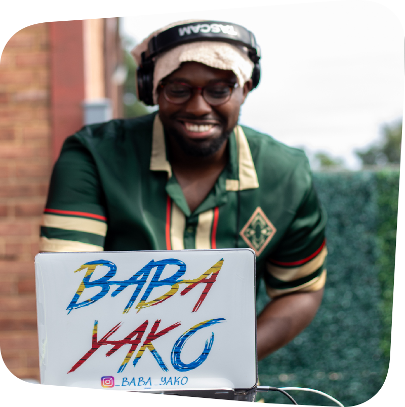 Smiling DJ Baba Yako standing behind a white laptop that displays his name in blue, red, and yellow letters. DJ Baba Yako wears headphones on his head and a green shirt.
