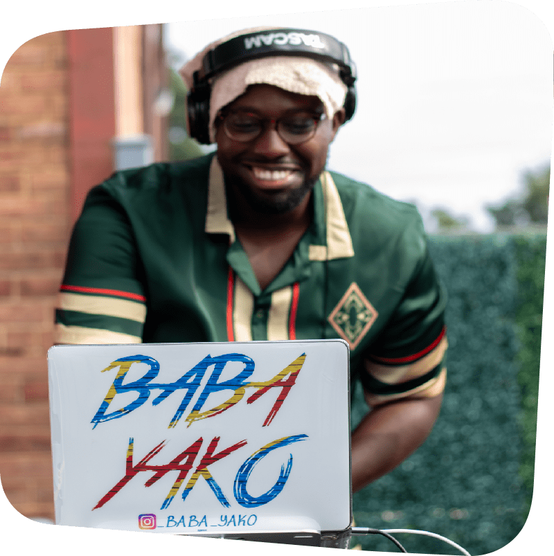 Smiling DJ Baba Yako standing behind a white laptop that displays his name in blue, red, and yellow letters. DJ Baba Yako wears headphones on his head and a green shirt.