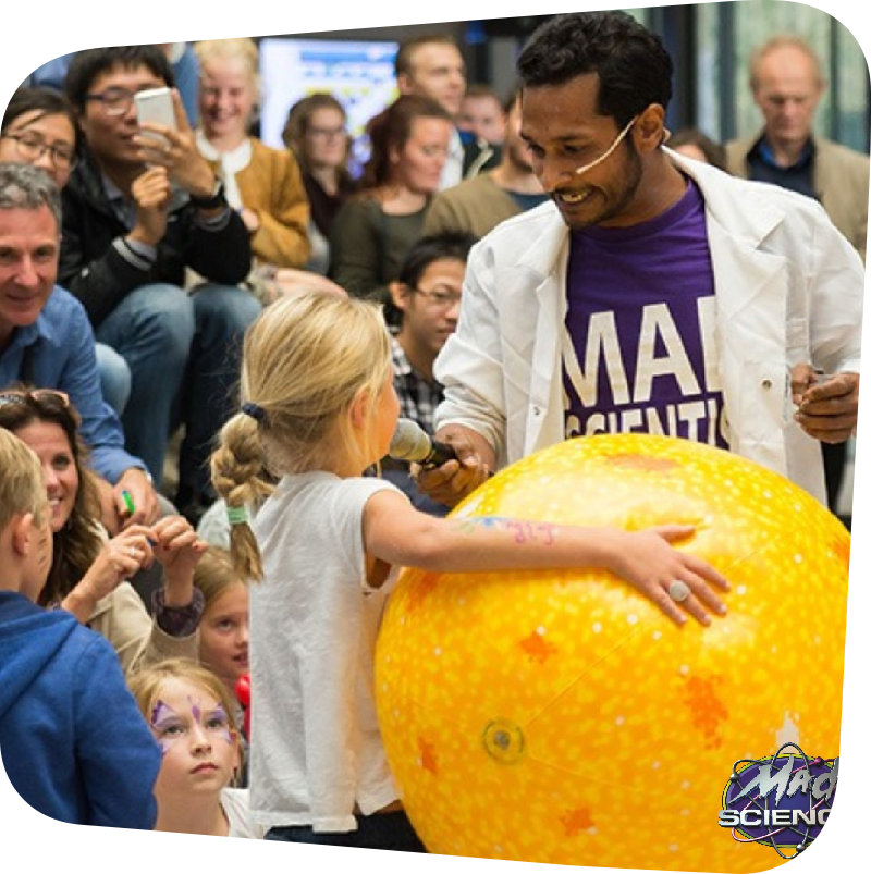 Foreground: Man wearing a lab coat holds microphone up to a young girl, who has her hand on top of a large yellow ball. Background: audience of children and guardians spectate.