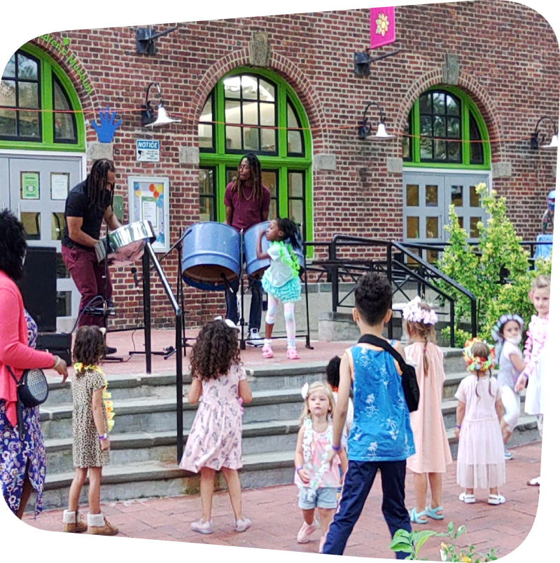 At the front of the outside of the museum: Standing on the top stair landing, two performers with steel drums and a little girl dancing. At the bottom of the landing: various children dancing.