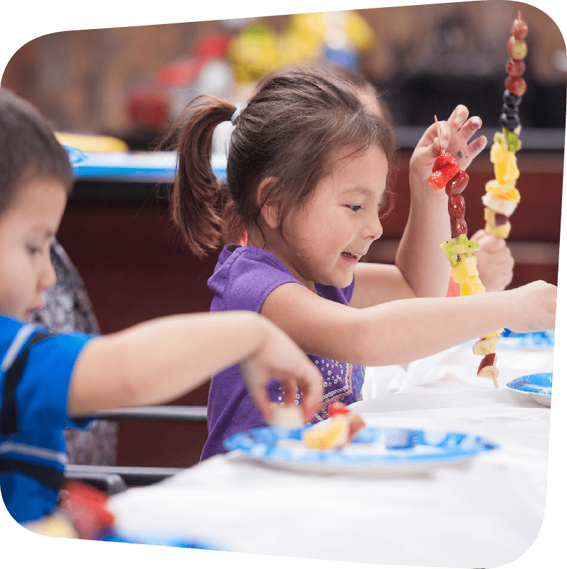 Two young children seated at table putting fruit and stick skewers. Smiling seated girl is central focus of image.