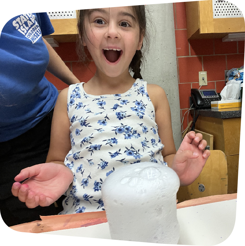 Girl smiling with open-mouthed amazement directly at viewer as in front of her on a table, bubbles froth out of a clear container in a chemical reaction.