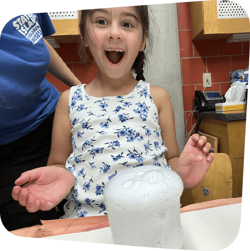 Girl smiling with open-mouthed amazement directly at viewer as in front of her on a table, bubbles froth out of a clear container in a chemical reaction.