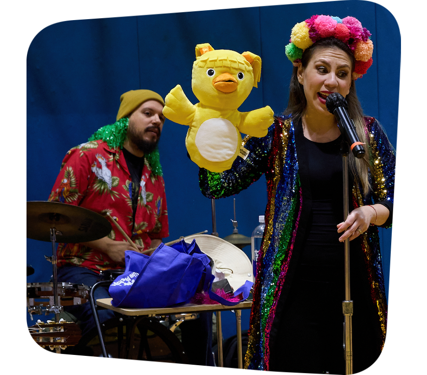 Right Foreground: A woman wearing a dress and colorful headpiece speaking or singing into a microphone with a yellow bird puppet on her hand. Left background: a bearded man in a red shirt sits behind a drum set.