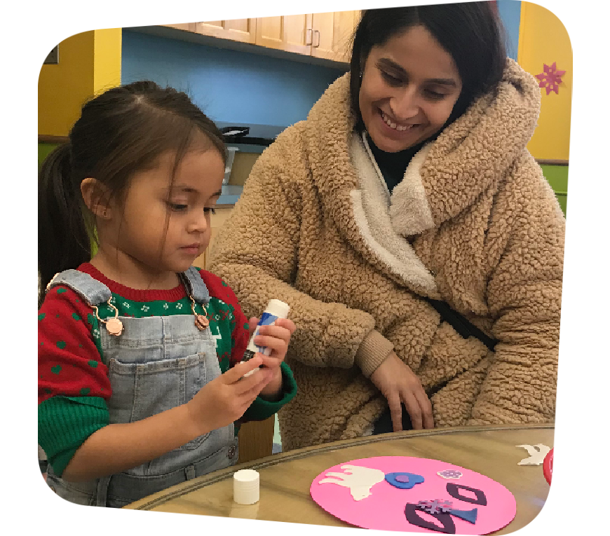A mother in a brown teddy-bear textured jacket smiles down at her daughter as the little girl focuses on twisting a glue stick. On the table they sit in front of is a pink circle decorated with white, blue, and purple cut-outs.