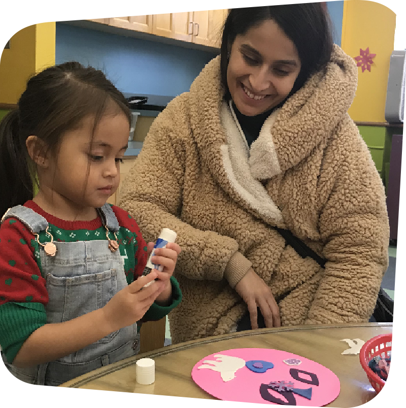 A mother in a brown teddy-bear textured jacket smiles down at her daughter as the little girl focuses on twisting a glue stick. On the table they sit in front of is a pink circle decorated with white, blue, and purple cut-outs.