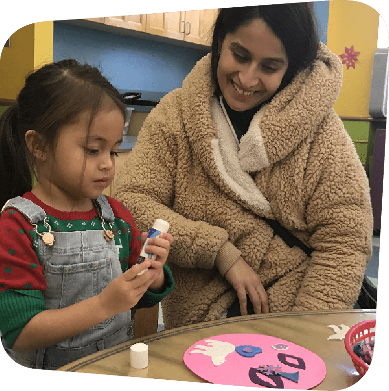 A mother in a brown teddy-bear textured jacket smiles down at her daughter as the little girl focuses on twisting a glue stick. On the table they sit in front of is a pink circle decorated with white, blue, and purple cut-outs.