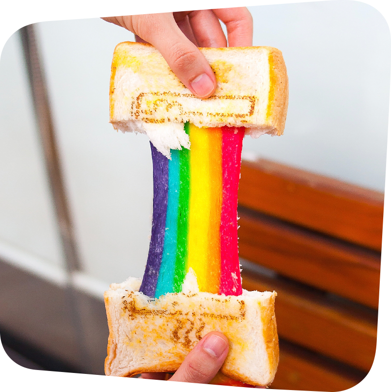 A pair of hands pulling apart two halves of a grilled cheese sandwich. The cheese stretches, the purple, blue, green, yellow, orange, and red of a rainbow.