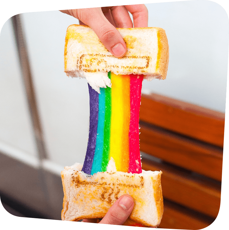 A pair of hands pulling apart two halves of a grilled cheese sandwich. The cheese stretches, the purple, blue, green, yellow, orange, and red of a rainbow.