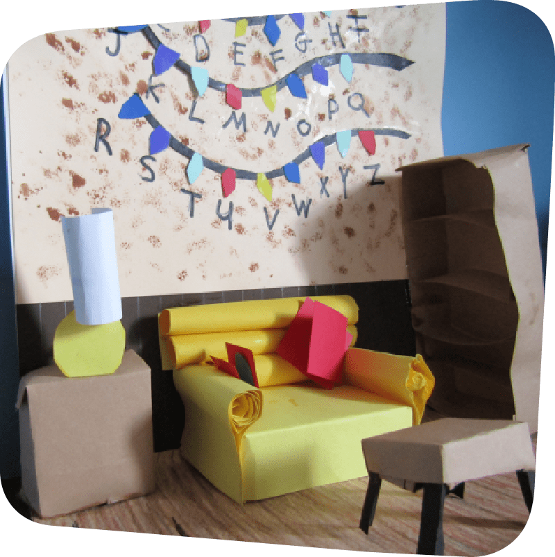 up-close of a mini model of the Walk-in Workshop craft. On a folded piece of paper, a multi-colored paper decoration hung on the wall with the letters of the alphabet. In front of the wall, a living room area with a table and lamp, beside which is a yellow couch, beside which is a brown bookcase, and a coffee table in front of the couch. These objects were folded to make them three-dimensional. The paper floor of model was decorated with a brown wood pattern.